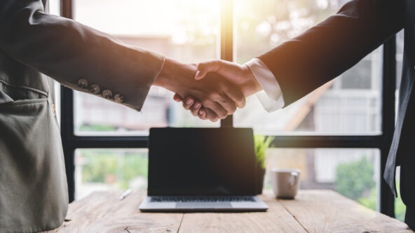 Two businessmen in suits shake hands in front of a laptop in a modern office.