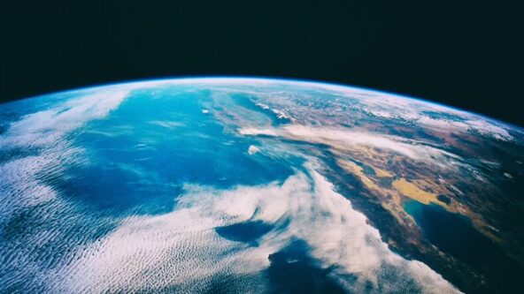 Blick auf die Erde aus dem Weltraum. Man sieht eine weite Fläche blauen Ozeans mit wirbelnden weißen Wolken. Auf der rechten Seite ist unter einem dunklen, weiten Himmel ein Stück Land zu sehen.